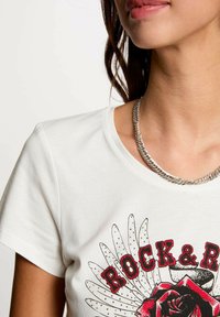 T-shirt in cotone bianco con una grafica floreale rossa e il testo "ROCK & ROLL", scollo rotondo e maniche corte. Collana a catena argentata visibile.