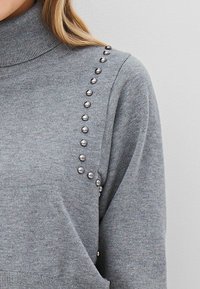 Pilkas megztinis su aukšta apykakle ir metaliniais diskelių akcentais ant vienos peties, išryškinantis švelnią tekstūrą ir prigludusią konstrukciją.