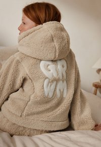 Barn med rødt hår iført en beige, fluffy hættejakke med hvide blokbogstaver "GR WM" på ryggen, siddende på en seng i et neutralt rum.