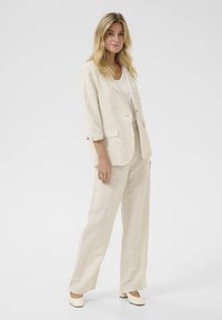 Beige linnen pak met een op maat gemaakt blazer en brede broekspijpen. Glad oppervlak, subtiele glans. Gekozen in combinatie met een licht topje en crèmekleurige schoenen.