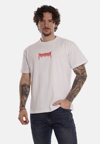 Maglietta di cotone bianca con maniche corte e scollo a girocollo. Presenta una scritta grafica rossa stampata sul petto. Indossata con jeans scuri.