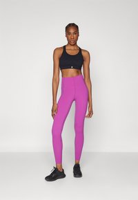 Schwarzer Sport-BH und hochtaillierte, enganliegende rosa Leggings. Das Material scheint dehnbar zu sein. Das Model trägt schwarze Sportschuhe. Neutrale Hintergrund.