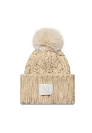 Strickmütze in hellbeige mit einem Zopfmuster, gekrönt mit einem Kunstfell-Pompon, mit einem weißen Logo-Patch am umgeschlagenen Bündchen.