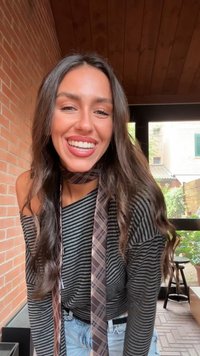 Maglietta a maniche lunghe a righe nera e grigia, abbinata a una cravatta marrone a motif. Jeans azzurri e onde morbide nei capelli scuri. Sorriso luminoso.