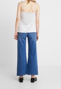 Camisola bianca con spalline sottili e regolabili, abbinata a jeans a gamba larga blu. I jeans presentano una trama sbiadita e dettagli sulle tasche posteriori.