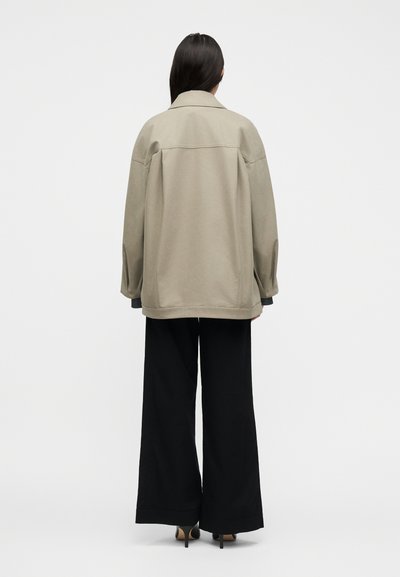 Άτομο με μακριά σκούρα μαλλιά που φορά χαλαρό μπεζ σακάκι, μαύρο παντελόνι φαρδύ και μαύρα παπούτσια με τακούνι, στέκεται με την πλάτη προς τα εμπρός σε λευκό φόντο.