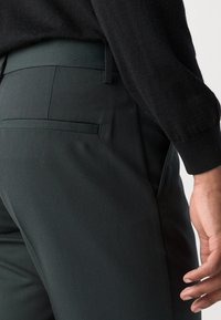 Donkergroene op maat gemaakte broek met een gladde textuur, voorzien van een achterzak, en gecombineerd met een zwarte trui. Het zijaanzicht toont de pasvorm en snit.