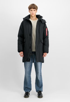 Un jeune homme portant un long manteau rembourré noir avec une étiquette rouge, une chemise beige, un jeans bleu et des chaussures noires, se tenant devant un fond blanc.