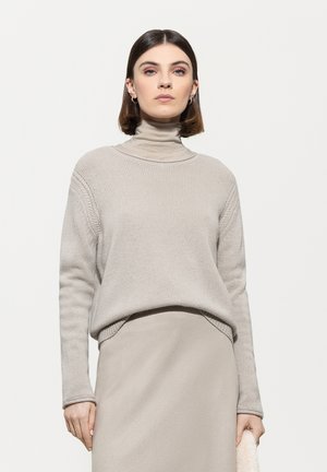 Beige Rollkragenpullover mit geripptem Struktur, lockerer Passform und detaillierter Stickerei. Kombiniert mit einem passenden Rock, beide in neutralen Farbtönen.