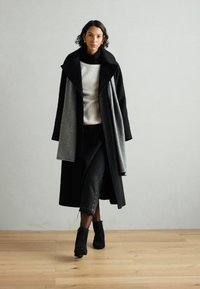 Abrigo largo negro y gris con un cuello sobredimensionado, sobre un suéter blanco de canalé, combinado con jeans negros y botas de tobillo. Suelo de madera, fondo neutro.