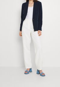 Kvinde iført navy blazer, hvid top med blonderkant, hvide bukser med høj talje og blå sandaler med blokhæl stående på hvidt gulv.