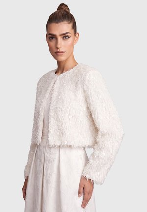 Woman with hair tied up wears a textured white cropped jacket over a light floral dress, facing forward with a neutral expression.