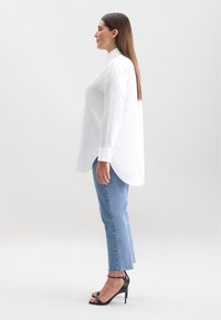Weißes langärmliges Hemd mit Knopfverschluss und asymmetrischem Saum, kombiniert mit hellblauen geraden Jeans und schwarzen Absatzschuhen mit Riemchen.
