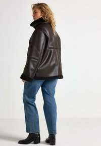 Brun læderjakke med en shearlingkrave, brede ærmer og en afslappet pasform, parret med blå denimjeans og sorte ankelstøvler.