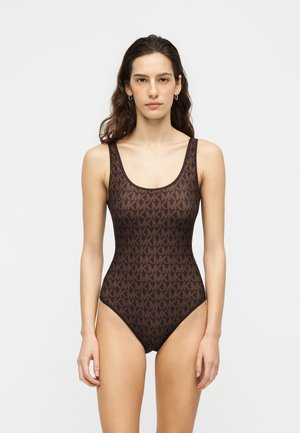 Femeie cu păr lung și închis la culoare poartă un body maro fără mâneci cu un model repetat de logo-uri MK, stând în fața unui fundal simplu și deschis.