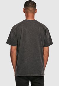 Hombre con cabello corto, vistiendo una camiseta de manga corta negra desgastada y pantalones negros, visto desde atrás contra un fondo blanco liso.
