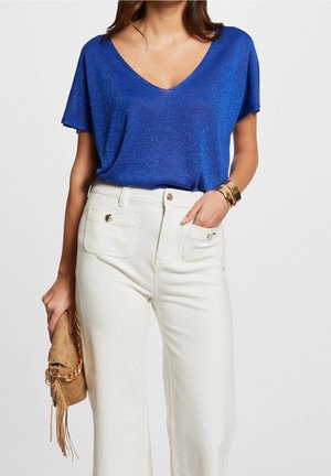 Kobieta ubrana w błyszczącą niebieską bluzkę z dekoltem w serek, białe spodnie z wysokim stanem z przednimi kieszeniami, złote bransoletki i trzymająca beżową torebkę z frędzlami.