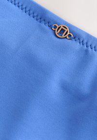 Gros plan d'un tissu bleu avec une couture en zigzag et un petit emblème métallique doré circulaire fixé près du bord.