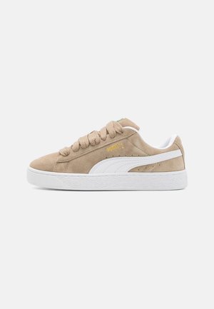 Beige ruskind Puma sneaker med hvid sål, hvid formstribe på siden, beige snørebånd og guld Puma-logo nær toppen af snørebåndsområdet.