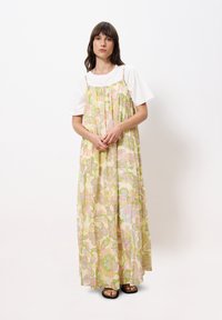 Maxikleid mit floralem Muster in Pastellfarben, verstellbaren Trägern und lockerer Passform, getragen über einem weißen Kurzarmhemd. Flache Sandalen.