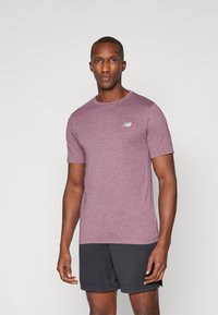Chemise à manches courtes en tissu violet chiné avec un col rond et un petit logo blanc sur la poitrine gauche. Portée avec un short de sport noir.
