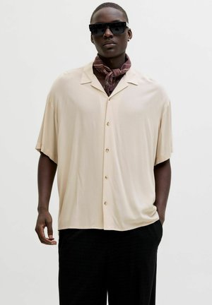 Mand iført oversize beige kortærmet skjorte med knapper, sorte bukser, mørke solbriller og et mønstret tørklæde bundet om halsen.