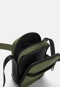 Kompakt grøn og sort taske med flere lynlåsecompartments, holdbart stof, justerbar rem og logomærke synligt indeni.