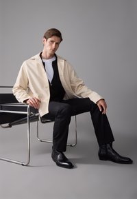 Chaqueta beige de botones sobre un abrigo negro con cremallera, combinada con pantalones negros y botas de cuero hasta el tobillo. Modelo sentado en una silla de metal.