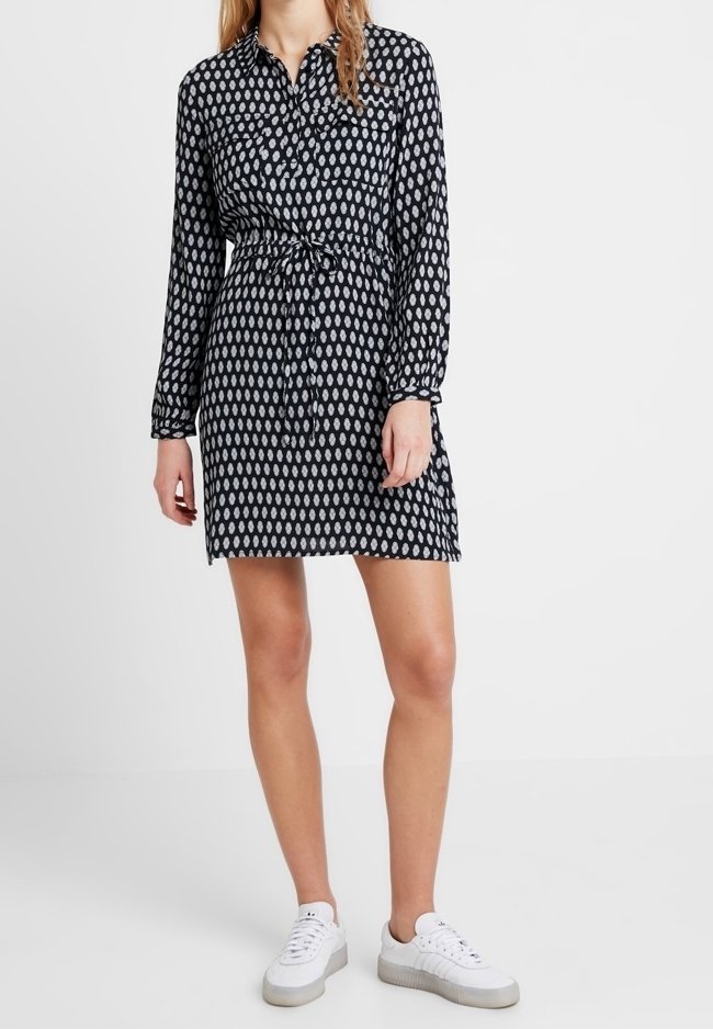 Femme portant une robe à manches longues à motif noir et blanc avec une ceinture à la taille, associée à des baskets blanches, se tenant sur un sol blanc.