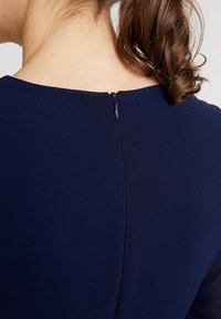 Vue de dos d'une personne portant une robe bleu marine avec une petite fermeture éclair dans le haut du dos et des cheveux bruns attachés lâchement.