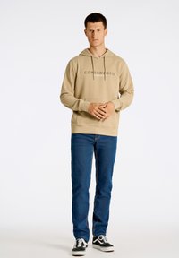 Felpa beige con cappuccio e cordino, logo "COPENHAGEN" centrato, tasca frontale. Abbinata a jeans blu e sneakers in bianco e nero.