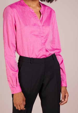 Kobieta w jasnoróżowej bluzce z długim rękawem i dekoltem w serek oraz w czarnych, wysokowiejskich spodniach na jednolitym tle.