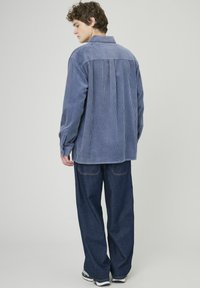 Chemise en velours côtelé bleu avec un design à pli dans le dos, manches longues et col. Associée à un pantalon en denim large bleu foncé.