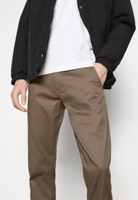 Veste noire zippée avec poignets élastiques, t-shirt blanc, et pantalon marron à finition lisse. Le pantalon possède une poche avant et une couture discrète.