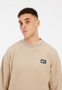 Felpa in pile beige con colletto a coste. Presenta una patch con il logo "PROTEST" nero e colorato sul lato sinistro del petto. Tessuto morbido.