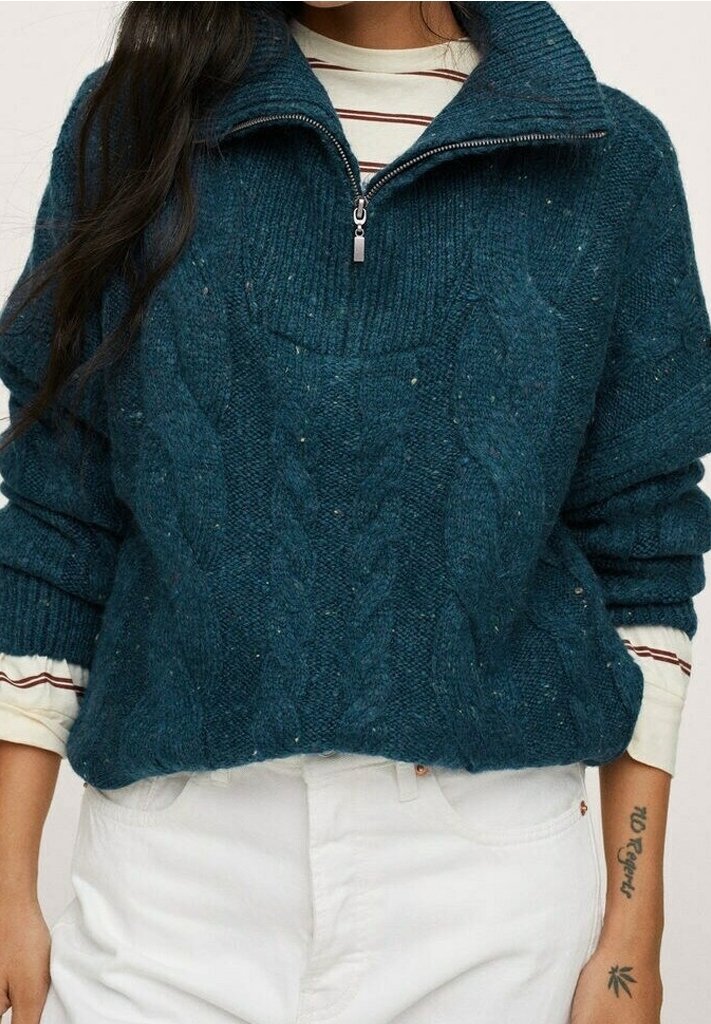 Femme portant un pull à demi-fermeture éclair en maille torsadée bleu sarcelle foncé sur une chemise à manches longues blanche et rouge rayée, avec un pantalon blanc.