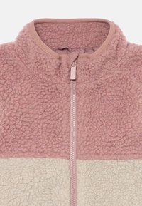 Fleecejacke mit hohem Kragen und Reißverschluss. Oberer Teil in sanftem Rosa und unterer Teil in Creme mit einer strukturierten, flauschigen Oberfläche.