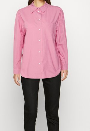 Kobieta w luźnej, różowej, długorękawowej koszuli zapinanej na guziki z kołnierzem oraz w czarnych spodniach, stojąca na jednolitym tle.