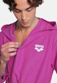 Accappatoio rosa con cappuccio e texture morbida, presenta un logo bianco "arena" sul petto, che evidenzia un design rilassato e casual.