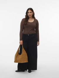 Frau mit langen dunklen Haaren, die ein braunes Langarmshirt und weite schwarze Hosen trägt und eine große beigetöne Wildleder-Shopper-Tasche hält.