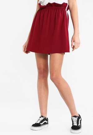 Woman wearing a short, high-waisted red skirt with a ruffled elastic waistband and black and white lace-up sneakers.