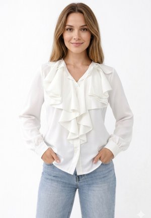 Jeune femme aux longs cheveux portant un chemisier blanc à volants et un jean bleu clair, les mains dans les poches, debout devant un fond blanc.