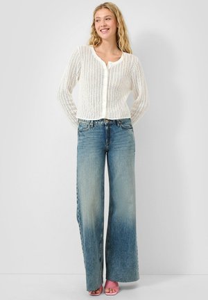 Lachtende vrouw met golvend blond haar draagt een witte gebreide vest met knopen, hooggetailleerde verwassen wijde jeans en roze open-teen hakken.