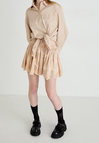 Beige checkered blouse tied at the waist, paired with a pleated, light beige skirt. Black platform shoes with straps, and knee-high black socks.