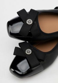 Bailarinas de charol negro con tiras de tela entrelazadas y botones decorativos de plata sobre un fondo blanco.
