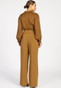 Vrouw met een knot in haar haar, gekleed in een bruine blouse met lange mouwen en hoge taille wijde broek, staand met haar gezicht naar rechts op een witte achtergrond.