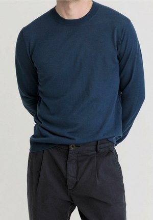 Mand iført en marineblå trøje med lange ærmer og mørke bukser, stående med hænderne bag ryggen mod en ensfarvet baggrund.