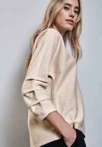Maglione beige chiaro con un motivo testurizzato, dotato di maniche a tre quarti e una vestibilità rilassata, indossato sopra dei pantaloni neri.
