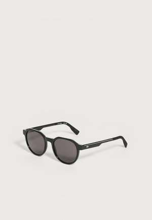 Lunettes de soleil rondes noires avec des verres foncés et des branches fines comportant un petit logo blanc sur le côté, posées sur une surface claire.