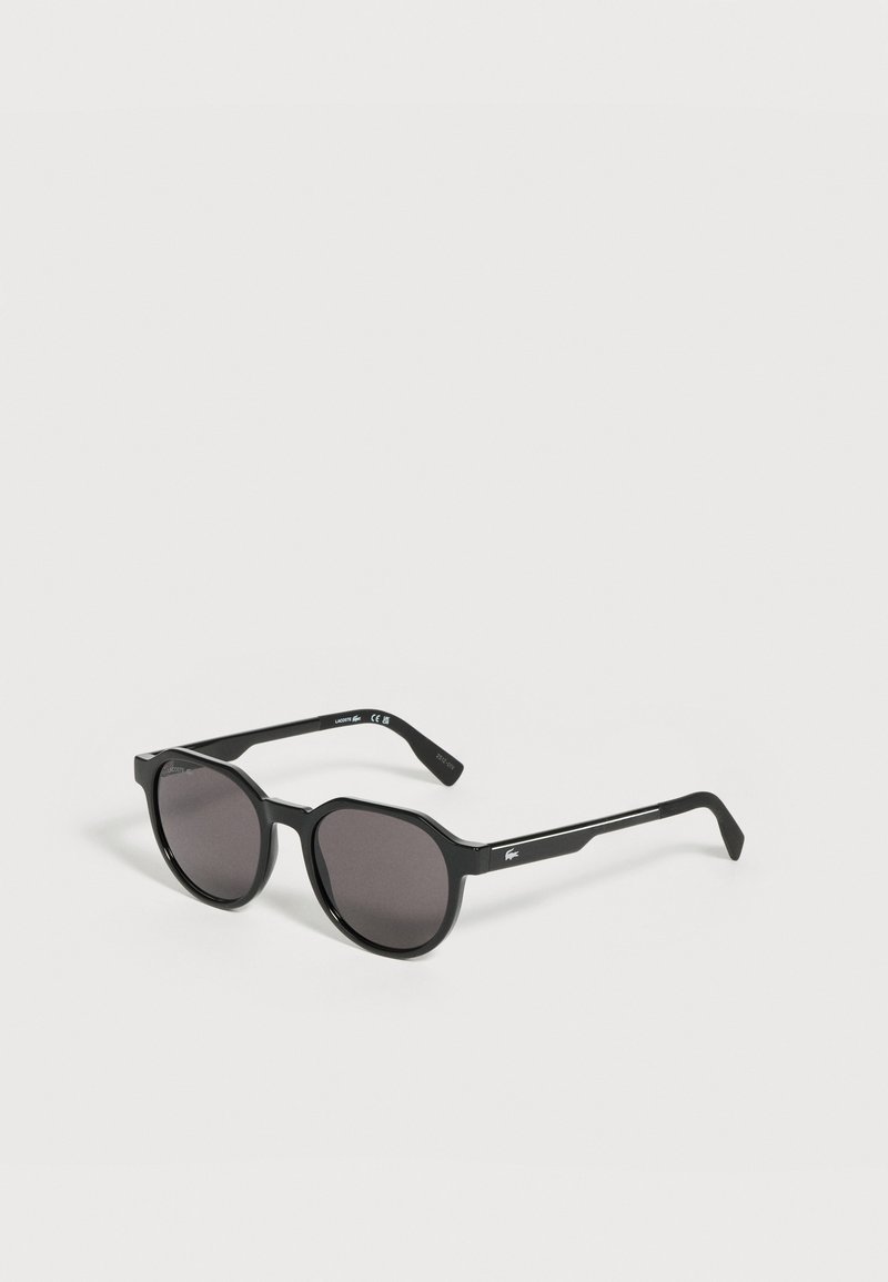 Lunettes de soleil rondes noires avec des verres foncés et des branches fines comportant un petit logo blanc sur le côté, posées sur une surface claire.